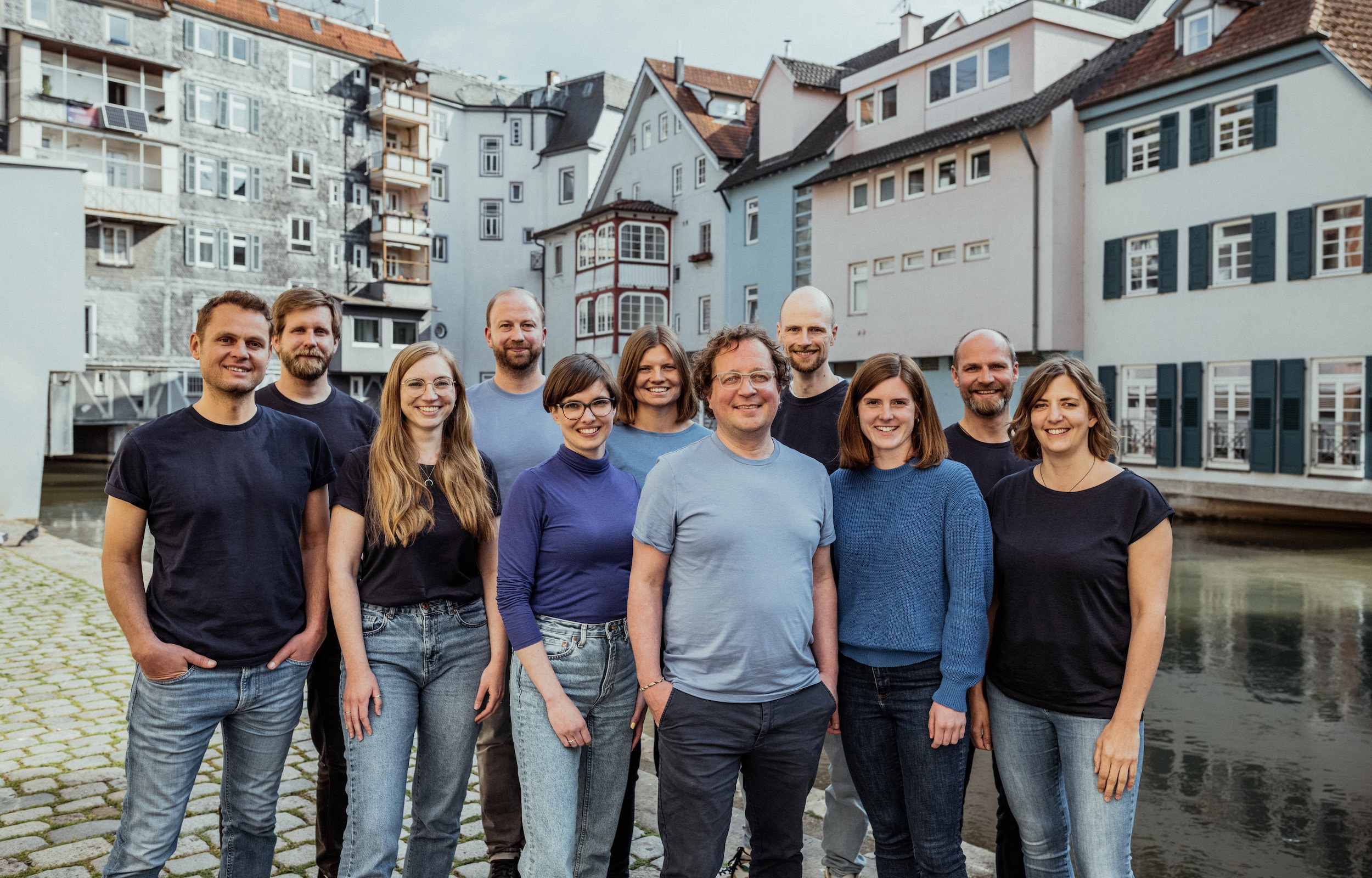 Foto mit dem Agenturteam, die 12 Personen lachen fröhlich, stehen als Gruppe am Ufer des Neckarkanals in der Esslinger Innenstadt, im Hintergrund ist das historische Häuserensemble, welches zu Esslingens Klein Venedig zählt